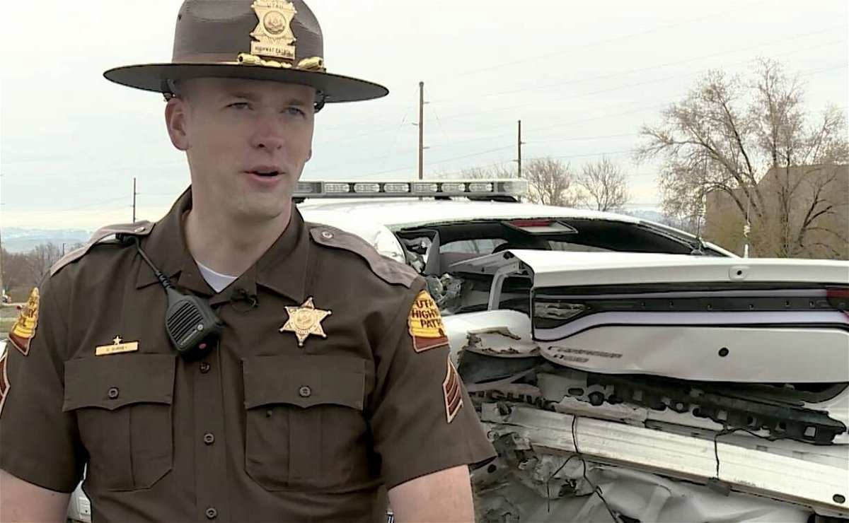 <i>KSL</i><br/>Utah Highway Patrol Sgt. Devyn Gurney speaks with members of the media on Thursday in front of his mangled patrol car. Gurney was struck by it in 2017 after it was rear-ended while he was conducting a traffic stop.