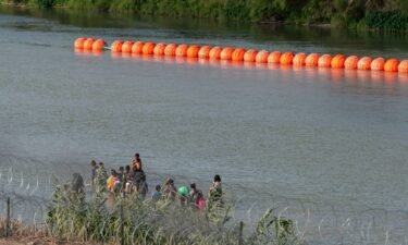 Migrants walk by a string of buoys placed on the water along the Rio Grande border with Mexico in Eagle Pass