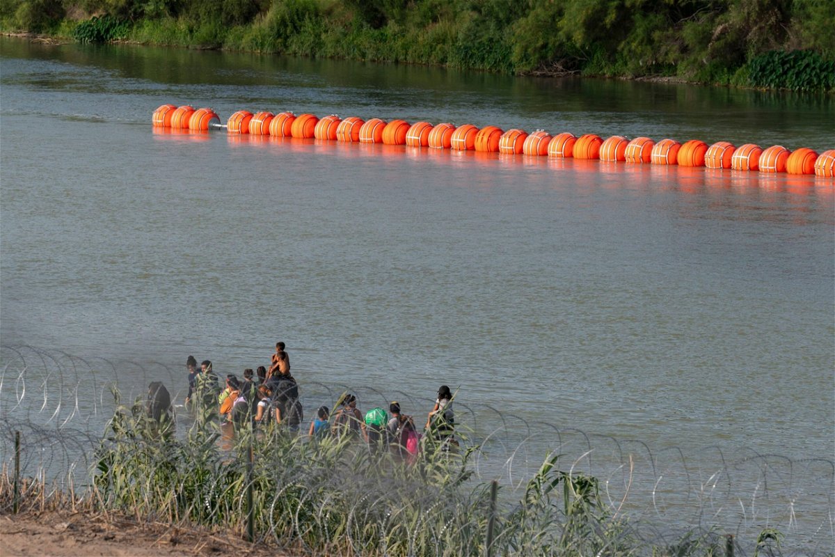 Migrants walk by a string of buoys placed on the water along the Rio Grande border with Mexico in Eagle Pass